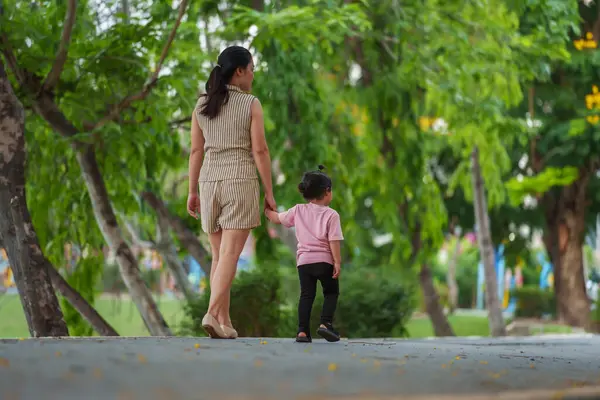 Yürüyen ve parktaki kaldırımda annesiyle el ele tutuşan küçük bir kız.