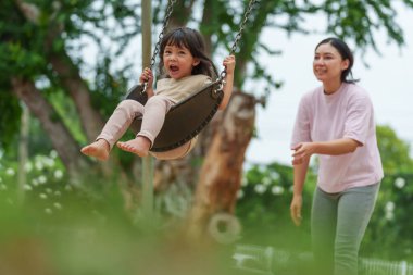 Neşeli küçük kız ve annesi açık hava çocuk parkında salıncakta oynuyor.
