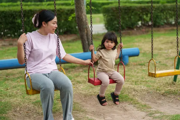 Neşeli küçük kız ve annesi açık hava çocuk parkında salıncakta oynuyor.