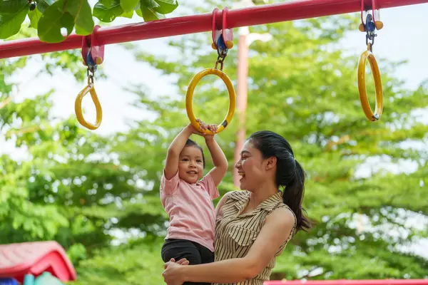 Anne kızını oyun sahasındaki yüzüklere asmak için tutuyor.