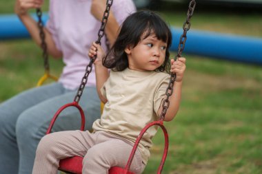Neşeli küçük kız ve annesi açık hava çocuk parkında salıncakta oynuyor.