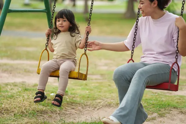Neşeli küçük kız ve annesi açık hava çocuk parkında salıncakta oynuyor.