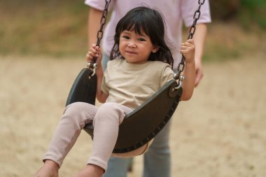 Neşeli küçük kız ve annesi açık hava çocuk parkında salıncakta oynuyor.