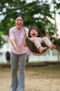 Neşeli küçük kız ve annesi açık hava çocuk parkında salıncakta oynuyor.