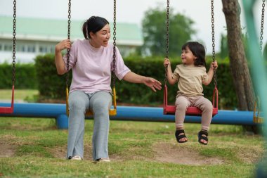Neşeli küçük kız ve annesi açık hava çocuk parkında salıncakta oynuyor.