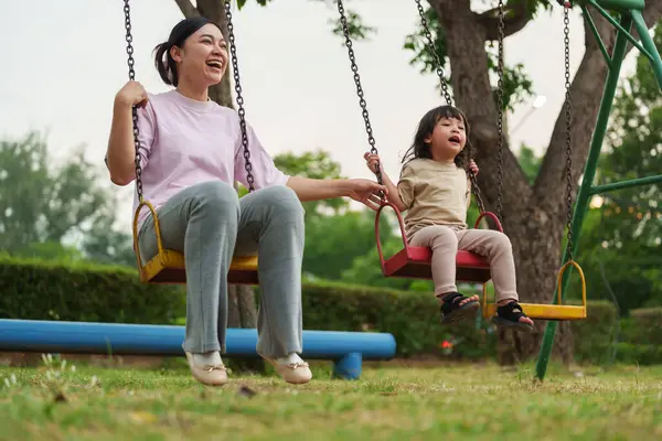 Neşeli küçük kız ve annesi açık hava çocuk parkında salıncakta oynuyor.