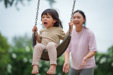 Neşeli küçük kız ve annesi açık hava çocuk parkında salıncakta oynuyor.