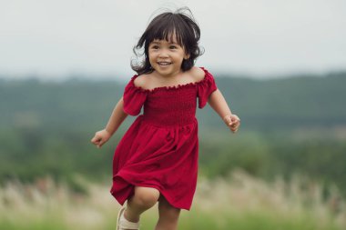 Kırmızı elbiseli mutlu kız çim tarlasında koşuyor.