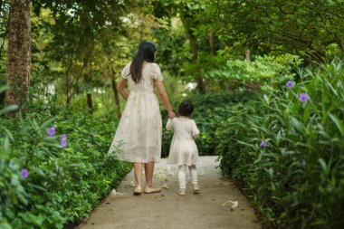 Çiçek bahçesinde yürüyen beyaz elbiseli anne ve küçük kız.