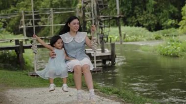 Mutlu anne ve küçük kız, doğada, dere kenarında salıncakta sallanıyor.