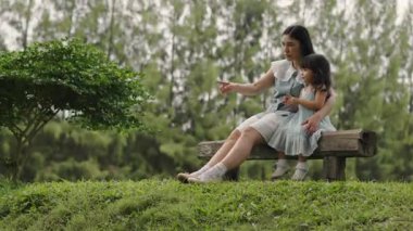 Mutlu anne ve küçük kız ahşap bir bankta oturuyor ve parkta birlikte konuşuyorlar.