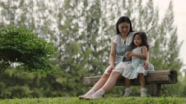 Mutlu anne ve küçük kız ahşap bir bankta oturuyor ve parkta birlikte konuşuyorlar.