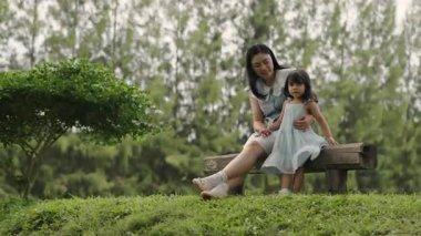 Mutlu anne ve küçük kız ahşap bir bankta oturuyor ve parkta birlikte konuşuyorlar.