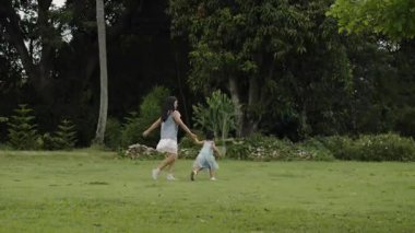 Anne, küçük kızın elini tutarken parkta çim tarlasında yürüme ve koşma oyunu oynuyor.