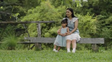Mutlu anne ve küçük kız ahşap bir bankta oturuyor ve parkta birlikte konuşuyorlar.