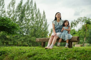 Mutlu anne ve küçük kız ahşap bankta oturuyor ve parkta birlikte konuşuyorlar.