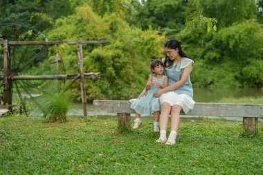 Mutlu anne ve küçük kız ahşap bankta oturuyor ve parkta birlikte konuşuyorlar.