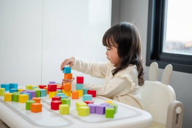 Küçük kız bir masanın üzerine küp inşa etmek için blok oyuncağı oynuyor.