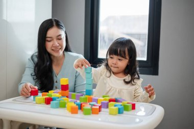 Yeni yürümeye başlayan mutlu bir kız annesiyle masada tahta blok oyuncakları oynuyor.