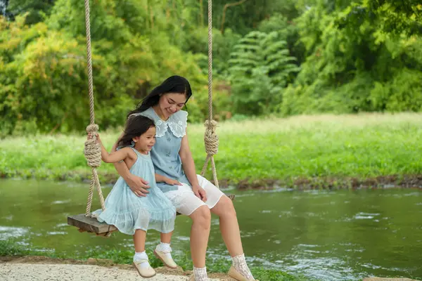 Mutlu anne ve küçük kız, doğada, dere kenarında bir ağaçta salıncakta sallanıyor.