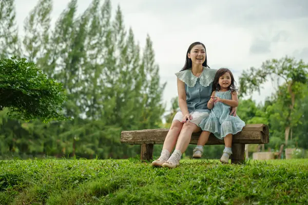 Mutlu anne ve küçük kız ahşap bankta oturuyor ve parkta birlikte konuşuyorlar.