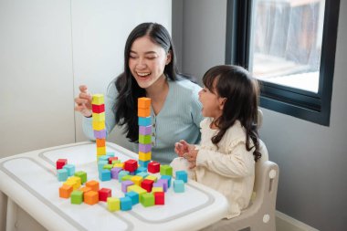 Yeni yürümeye başlayan mutlu bir kız annesiyle masada tahta blok oyuncakları oynuyor.