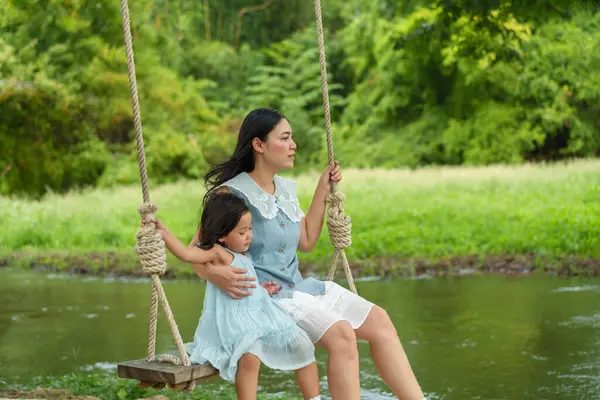 Mutlu anne ve küçük kız, doğada, dere kenarında bir ağaçta salıncakta sallanıyor.