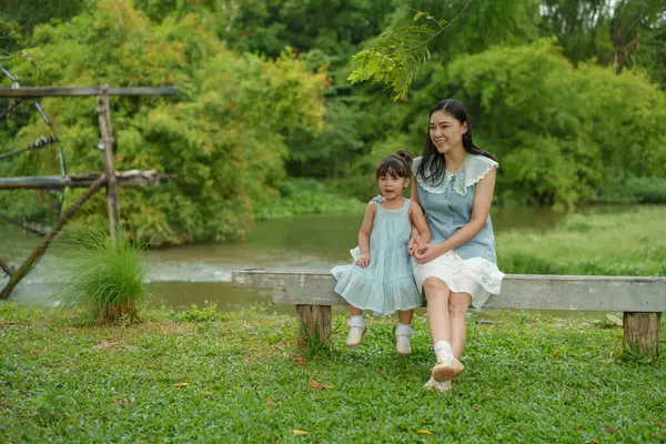 Mutlu anne ve küçük kız ahşap bankta oturuyor ve parkta birlikte konuşuyorlar.