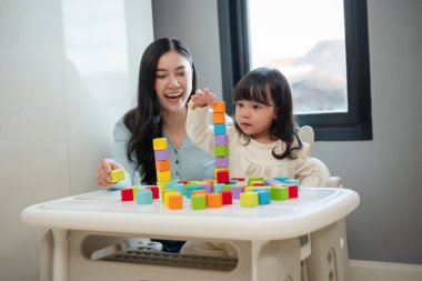 Yeni yürümeye başlayan mutlu bir kız annesiyle masada tahta blok oyuncakları oynuyor.
