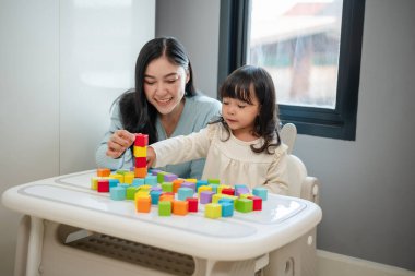 Yeni yürümeye başlayan mutlu bir kız annesiyle masada tahta blok oyuncakları oynuyor.