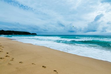 Bulutlu bir gökyüzünün altında kumlu bir plaja yuvarlanan dalgaların sakinleştirici güzelliğini hissedin..