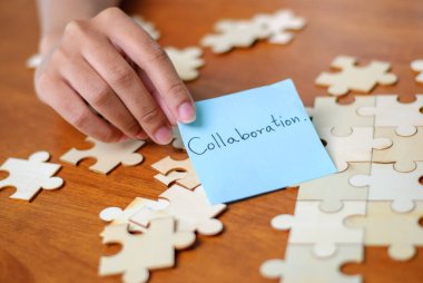 Bir el, dağınık yapboz parçalarının arasında iş birliği yaparak takım çalışmasını simgeleyen bir notu tutuyor..