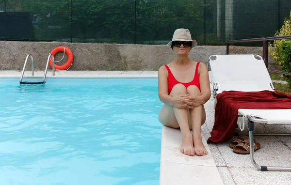 Middle-aged woman wearing a red bathing suit and a panama hat sitting by the edge of a swimming pool
