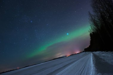 Snowy road leading through winter landscape. Stars and northern lights in the night sky.