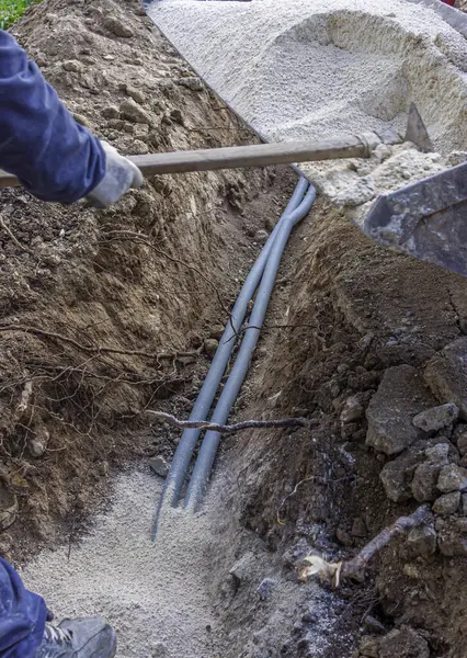 Worker covers electrical conduits with fine sand in a trench dug into the ground