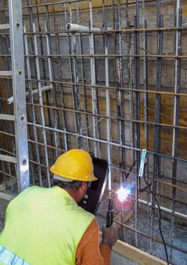Sarı kasket ve güvenlik yeleği giyen inşaat işçisi, kaynak işleminden kıvılcımlar görünen bir inşaat sahasında inşaat demirine kaynak yapıyor..