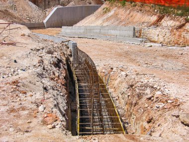 Formalite ve çelik inşaat ızgarası olan inşaat sahasının temelinde beton bir duvar, biçimlendirme ve çelik inşaat demirleri, kayalık ve kumlu toprakla çevrili..