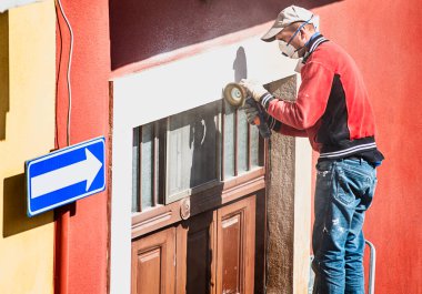 Kırmızı ceketli ve kotlu işçiler ahşap bir kapının üzerinde sürtünmek veya cilalamak için elektrikli bir alet kullanırlar.