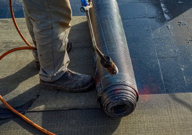 Zedelenmiş zarı olan su geçirmez çatı döşemesi. İşçinin ayaklarına ve zar yuvarlanmasına yakın çekim, başvuru sürecini gösteriyor. Fata Morgana etkisi