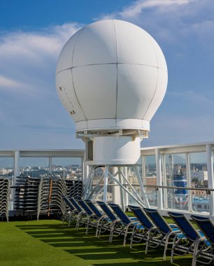 Bir yolcu gemisinin güvertesinde büyük beyaz bir uydu kubbesi. Yapay çimlerin üzerine istiflenmiş ve dizilmiş şezlonglar bulutlu bir gökyüzünün altında şehir manzaralı.