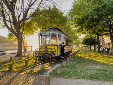 PORTO, PORTUGAL - CIRCA APRIL 2023: Porto Caddesi 'nde ilerleyen ahşap tarihi vintage sarı tramvay 203, şehrin sembolü. Massarelos 'ta gün batımında arka planda Arrabida köprüsü olan eski bir tramvay geçiyor. Portekiz. Io için vazgeçilmez ulaşım