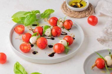 Domatesli şişler, mozarella topları, fesleğen ve baharatlar beyaz masada, yakın plan. İtalyan ev yapımı yemekler ve sağlıklı bir diyet konsepti..
