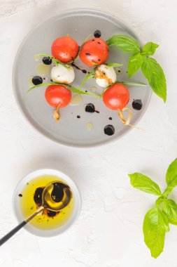 Domatesli şişler, mozarella topları, fesleğen ve baharatlar beyaz masada, yakın plan. İtalyan ev yapımı yemekler ve sağlıklı bir diyet konsepti..