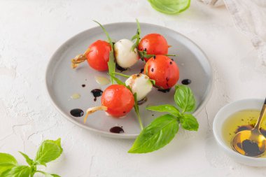 Domatesli şişler, mozarella topları, fesleğen ve baharatlar beyaz masada, yakın plan. İtalyan ev yapımı yemekler ve sağlıklı bir diyet konsepti..