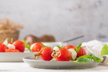 Domatesli şişler, mozarella topları, fesleğen ve baharatlar beyaz masada, yakın plan. İtalyan ev yapımı yemekler ve sağlıklı bir diyet konsepti..