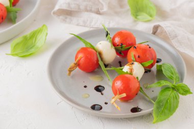 Domatesli şişler, mozarella topları, fesleğen ve baharatlar beyaz masada, yakın plan. İtalyan ev yapımı yemekler ve sağlıklı bir diyet konsepti..