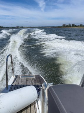 Bir sürat teknesi, açık, güneşli mavi gökyüzünün altında arkasında iz bırakarak suyun üzerine zum yapıyor. Sahne enerji ve heyecan dolu. Ria de Aveiro, Ovar - Portekiz.