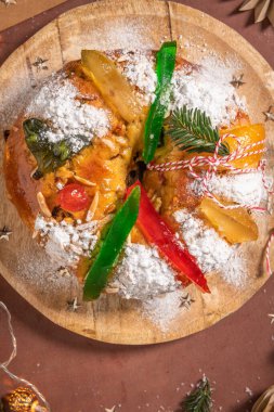 Colorful candied fruits and powdered sugar on a traditional Bolo Rei or Kings Cake, set on a wooden board amidst warm holiday decorations and lights. Is tradicional for Christmas in Portugal, Carnival or Mardi Gras. Top view clipart