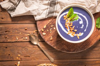 Bir kase canlı mor popato çorbası krema, kavrulmuş badem ve taze otlarla süslenmiş, ahşap bir tahtanın üzerinde dinleniyor..