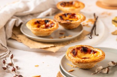 Portekiz kremalı tart koleksiyonu olan Pastel de Nata, yanında tarçın tozu, vanilya çekirdeği ve küçük altın kaşıklarla birlikte açık gri tabaklarda sergilenmektedir. Söğüt dalları bir bahar dokunuşu ekler.